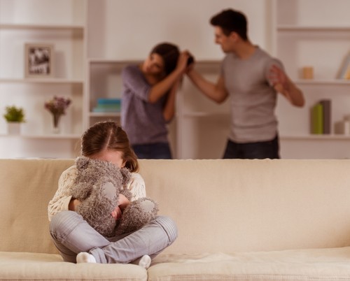 Young female child on sofa covering her face with a teddy bear as man pulls her mother's hair