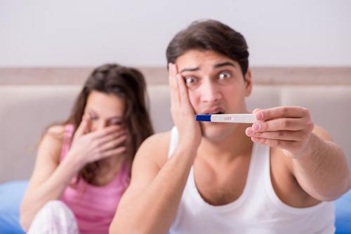 man and woman in bed. man is holding up a pregnancy test device and looking upset.