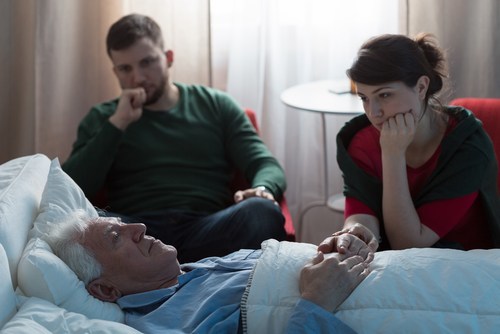 young man and woman sitting in hospital room worried about older man who is lying in the hospital bed