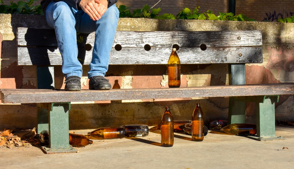 Persona sentada en un banco con varias botellas vacías de alcohol.