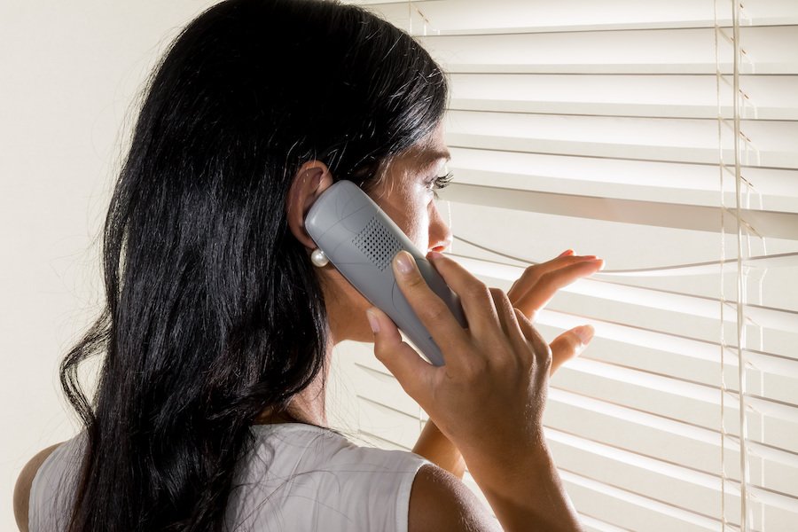 Woman on phone looking through window blinds at person