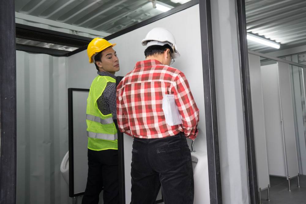 One man looking over the urinal stall in the direction of another man's stall.