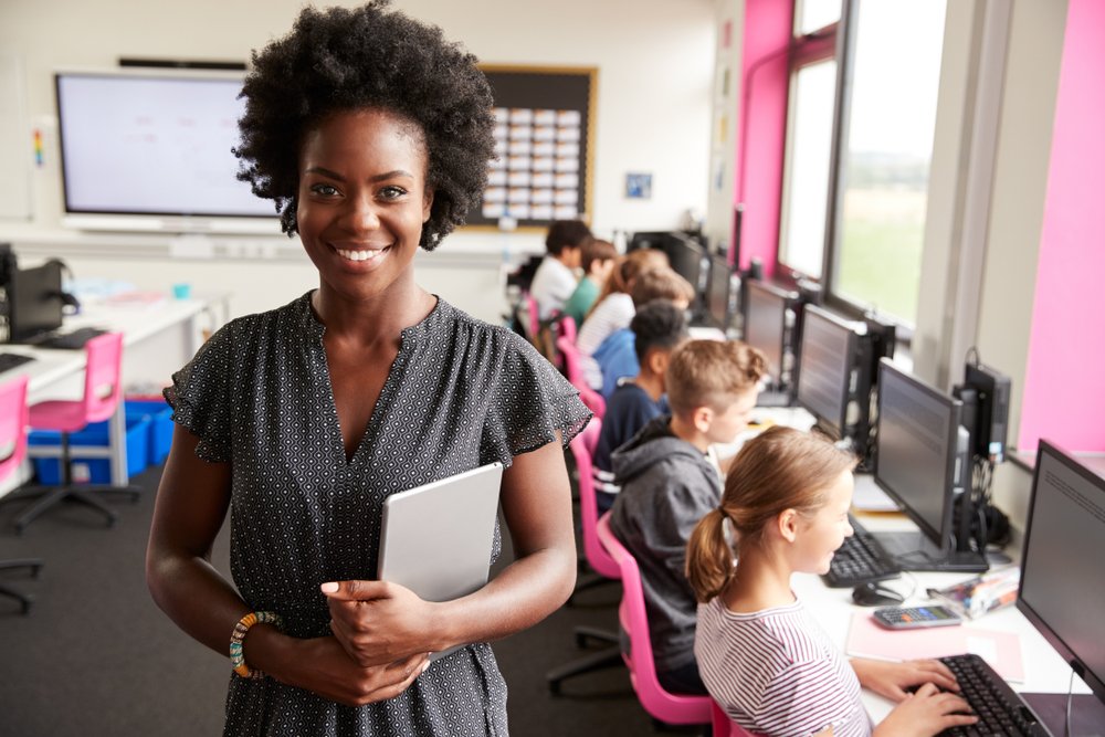 A smiling teacher in her classroom with her students behind her.