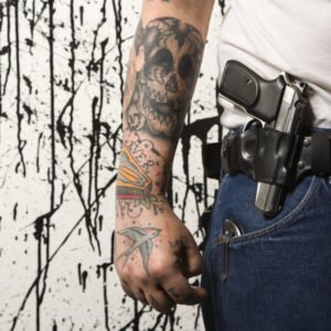 Closeup of man's belt holster carrying a firearm next to his tattooed arm.