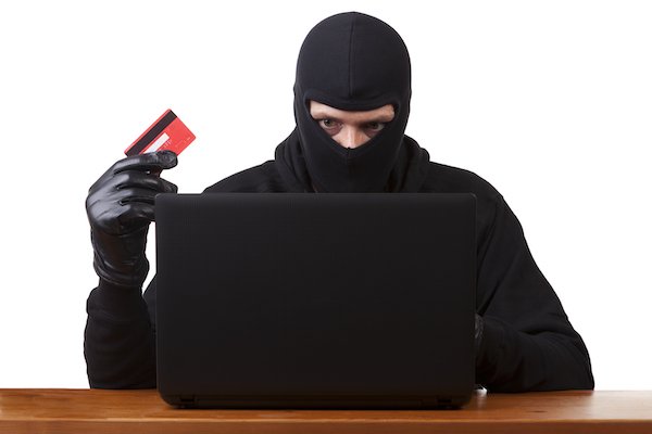 Ladrón con máscara detrás de una computadora portátil con una tarjeta de crédito robada