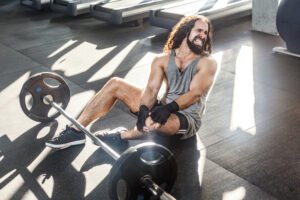 curly-haired young man with a beard winces in pain as he holds his knee, he is on the floor of a gym next to a straight barbell with 45 pound weights on each end