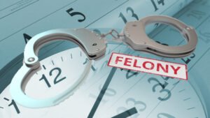 Composite collage image of handcuffs and the word "felony" in a red stamp, over top of a blended, half-opacity, image of an analog clock face with hands reading the time 3:08, over a calendar of black numbers representing days of an unknown month in an unknown year.
