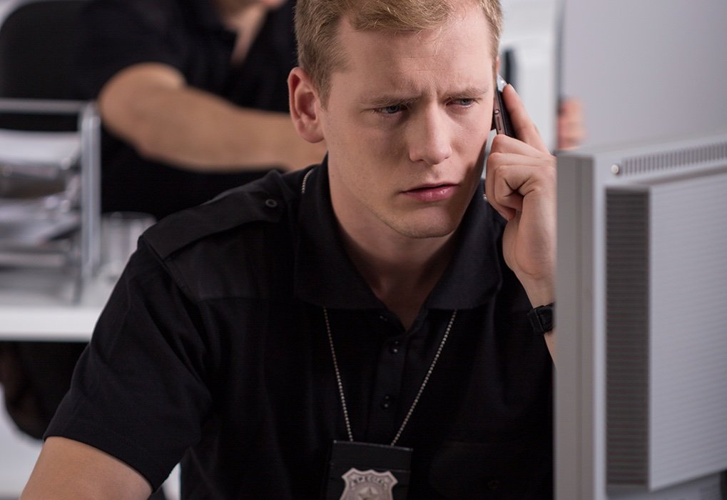 Young male police investigator sitting in a cubicle at the police station wears a badge around his neck as he talks on the phone and uses a computer