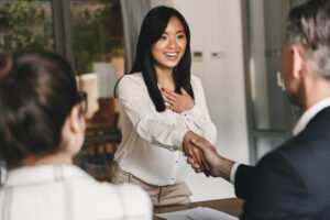 Interior of modern office. Young Asian woman in business attire, long sleeve white blouse and khaki pants, is smiling and overwhelmed with joy as she reaches across a desk to shake the hand of a Caucasian man with graying hair, who is wearing a business suit. Next to him sits a Caucasian woman with her dark brown hair in a bun, she is wearing thick-brimmed eye glasses.