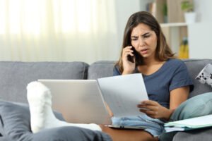 Mid 30's Caucasian woman wearing shorts and a t-shirt sits on a couch in a living room with her foot in a cast elevated on a footrest. Her brow is furrowed as she talks on the phone while scrutinizing paperwork that she holds in her hand. An open laptop computer sits on her legs in front of her.