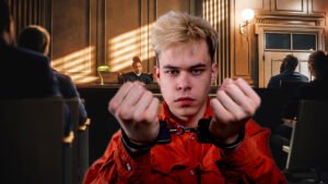 handcuffed juvenile teen in an orange jumpsuit stands in a courtroom with a judge and gallery.