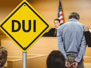 Man in handcuffs stands in front of a judge in a courtroom with his head lowered, there is an illustration of a yellow yeild street sign with the word "DUI" on it.